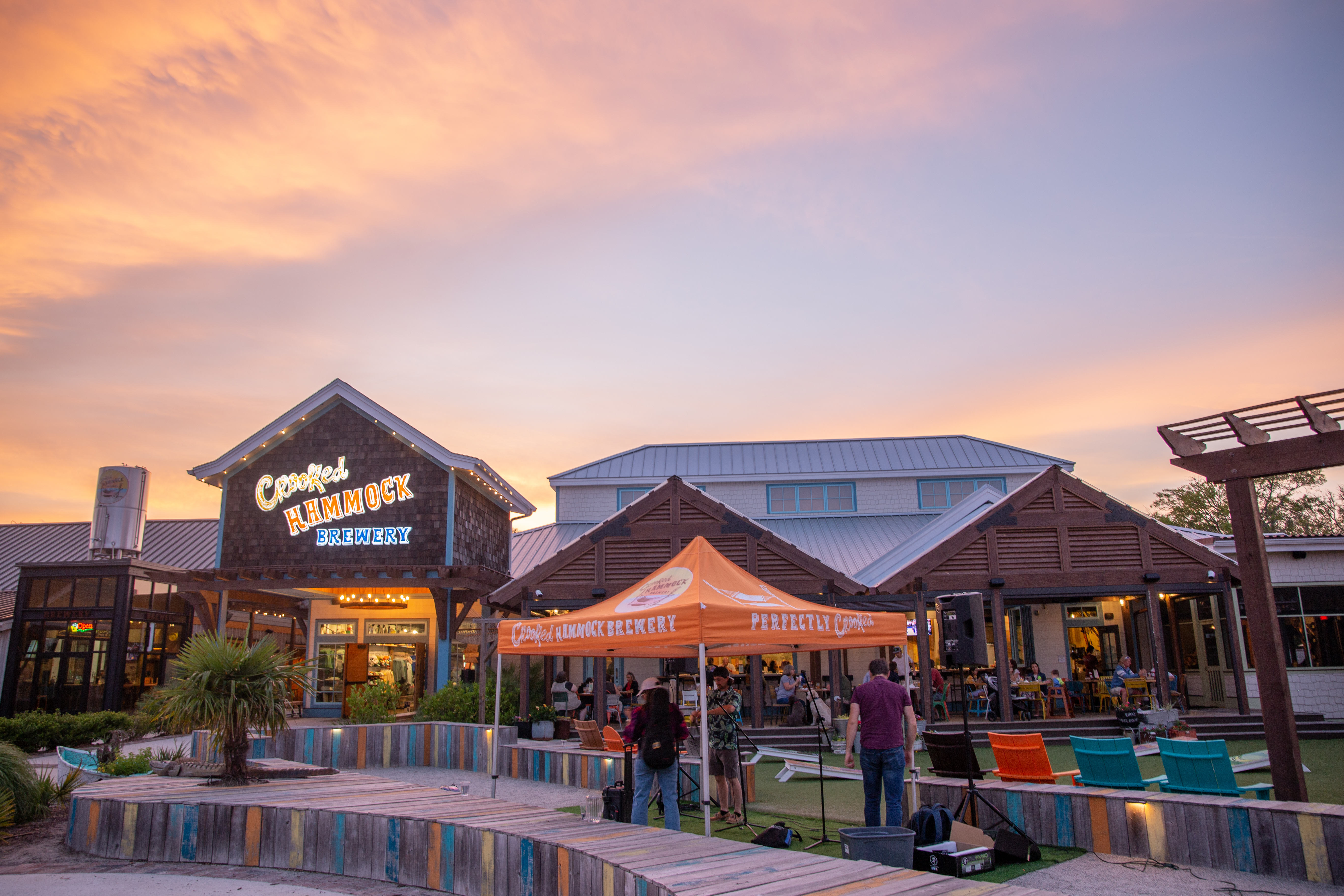 Crooked Hammock Brewery at sunset with outdoor event tent, colorful seating, and visitors enjoying craft beer and entertainment in a lively waterfront gathering space.