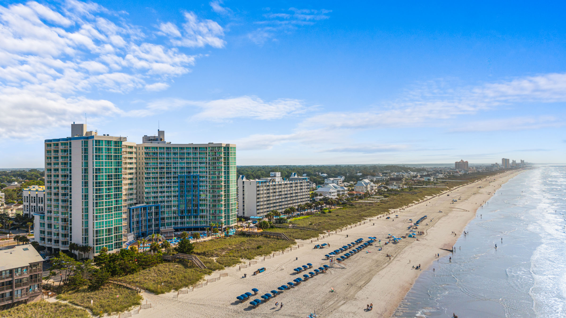 Colorful beachfront resort hotels overlooking a wide sandy beach with blue umbrellas, ocean waves, and distant city skyline on a sunny day.