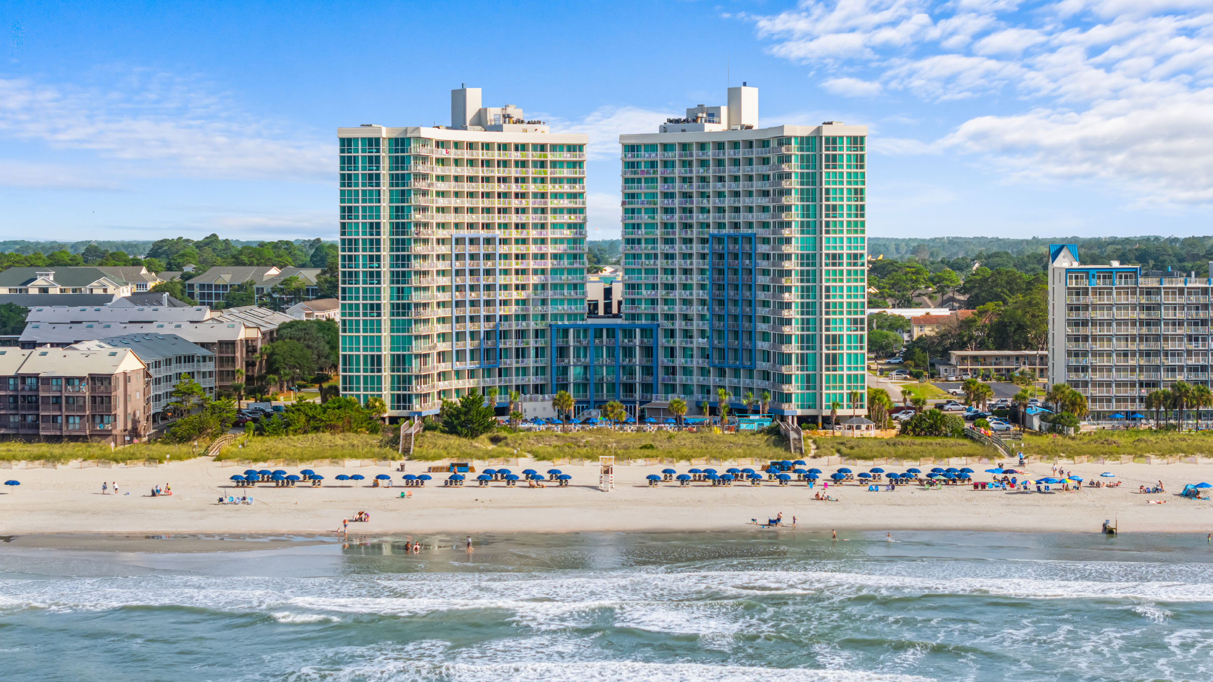 Beachfront resort hotels with blue umbrellas and sandy shores, ocean waves, and lush green landscape in background.