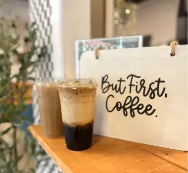 2 Coffees resting on a table next to a sign that reads "But first, coffee"