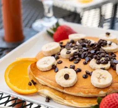 Fluffly banana pancakes topped with chocolate ships and surrounded by fruit on a plate.