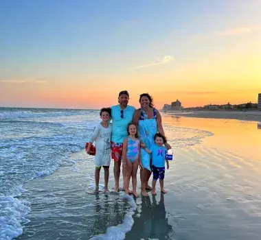 A family of 5 poses on the beach at sunset.
