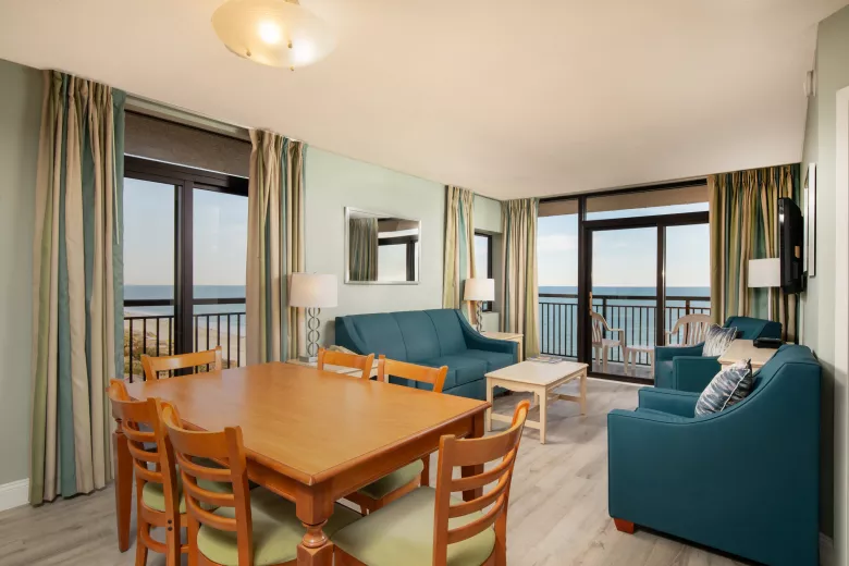 Living room with a wooden dining table with six chairs, two sofas, and floor to ceiling windows with ocean views