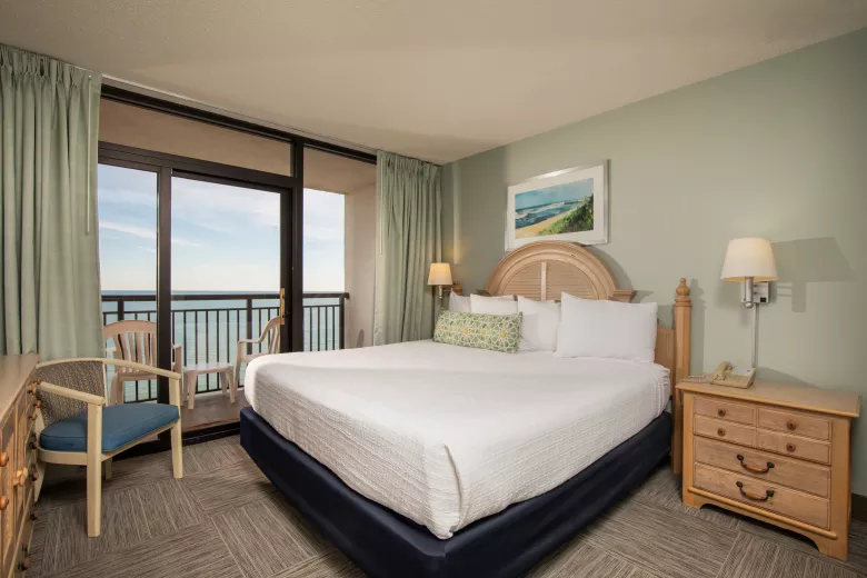 A bedroom with a king size bed, chair, dresser and nightstand with a large window overlooking the ocean