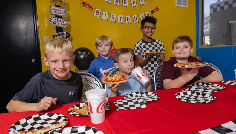 Kids enjoying pizza and drinks at a racing-themed birthday party with checkered decorations and Happy Birthday banner.