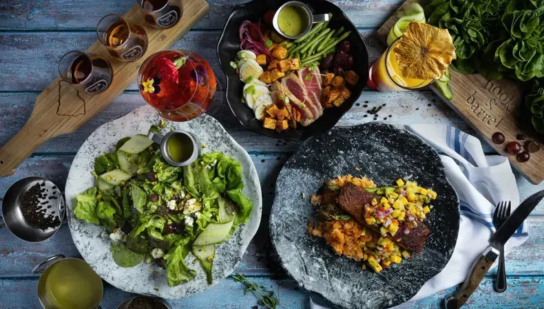 Artfully plated farm-to-table cuisine with fresh greens, grilled vegetables, and proteins paired with craft beverages on rustic blue wood.
